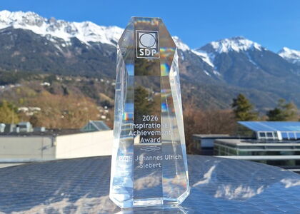 Glastrophäe des SDP Inspirational Achievement Award 2026 vor verschneitem Bergpanorama